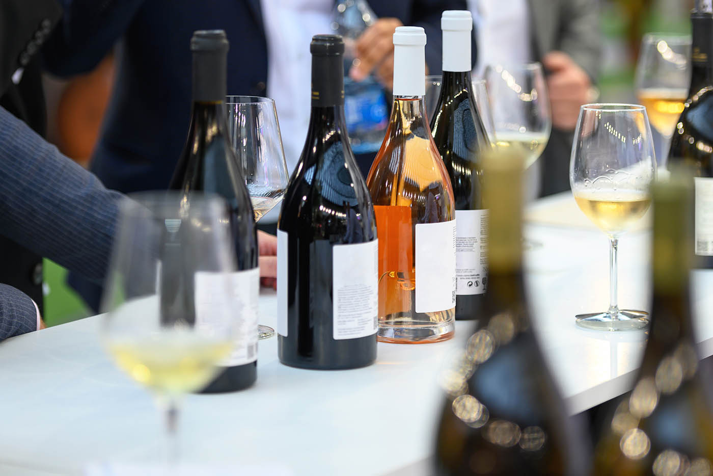 Various wine bottles and glasses filled with white and rosé wine sit on a white counter at a social event.