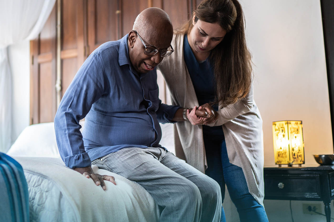 A young woman, a home health aide, supports an elderly Black man as he slowly stands up from the edge of his bed.
