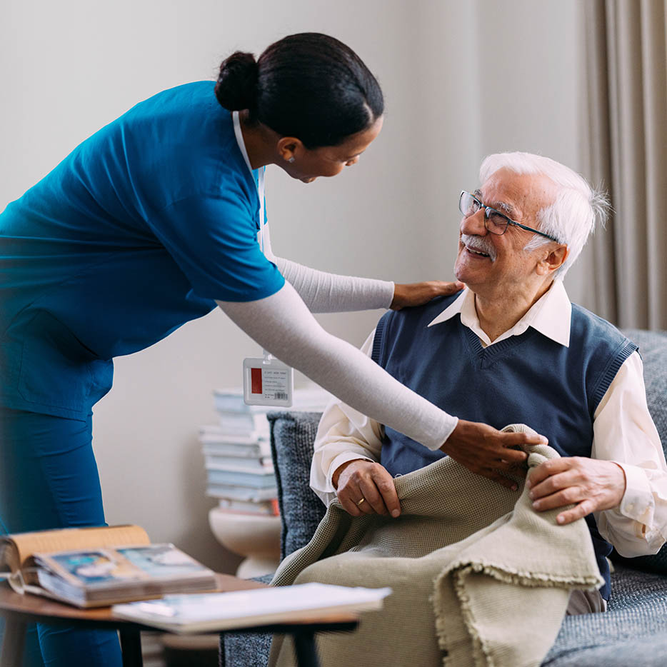 A smiling Black female home health aide in blue scrubs adjusts a blanket for a happy, elderly Caucasian man seated in a chair indoors.