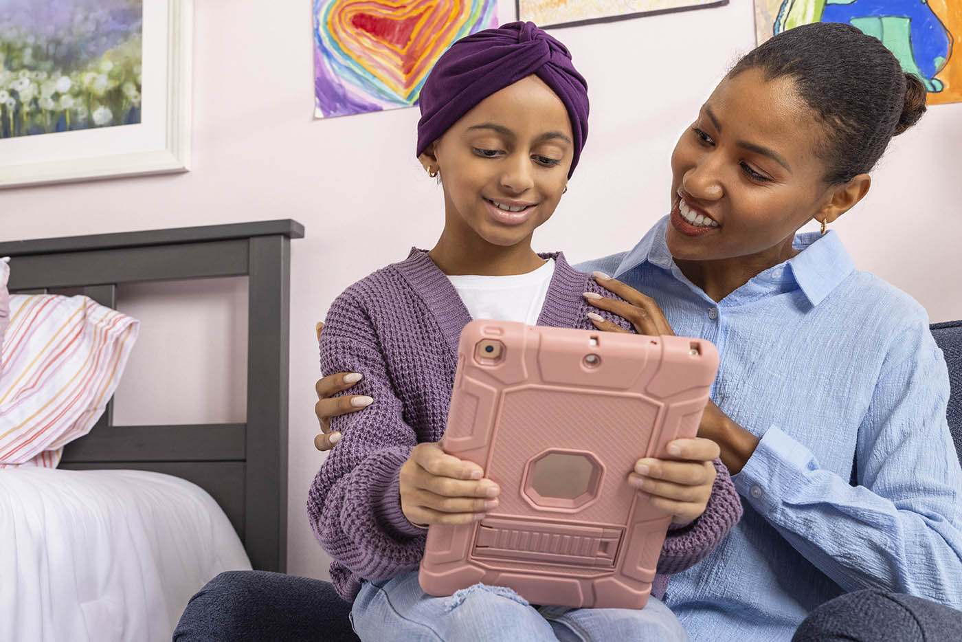 A smiling young girl in a purple headscarf holds a pink-cased tablet. Her mother, wearing a blue shirt, sits behind her, gently touches her shoulder.