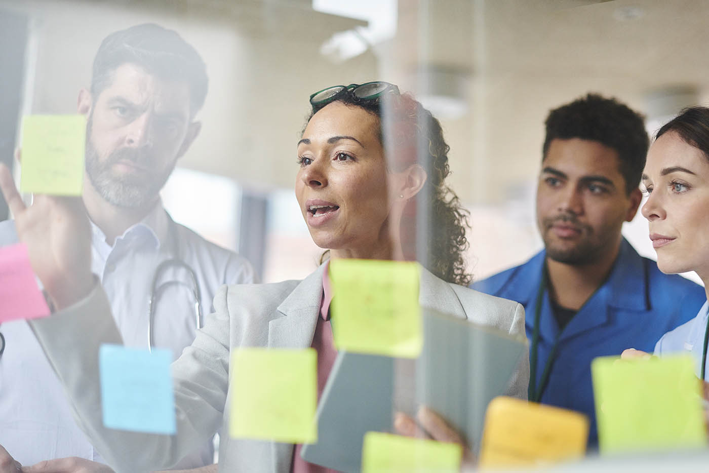 A diverse clinical and administrative team collaborates on a strategy, with a central woman talking near a clear board covered in sticky notes.