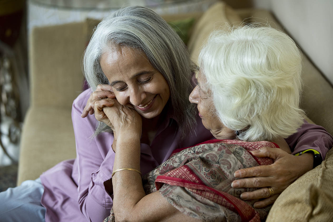 A smiling middle-aged woman with long gray hair embraces a white-haired older woman, leaning her cheek on their clasped hands.