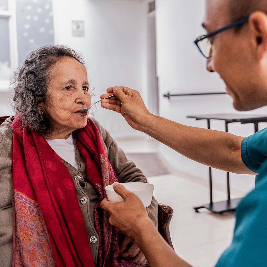 A nurse feeding a spoonful of food to an elderly woman sitting in a chair.