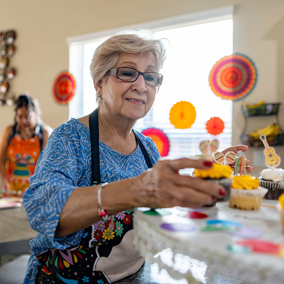 A smiling elderly woman wearing an apron and glasses arranges cupcakes with colorful decorations with a helper in the background.