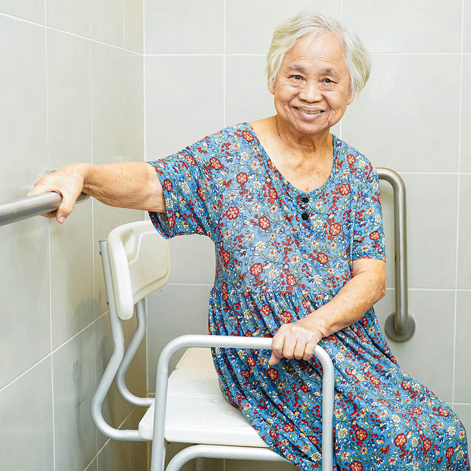 A smiling, elderly Asian woman sits on a white shower chair, safely holding a metal grab bar attached to the tiled wall behind her.