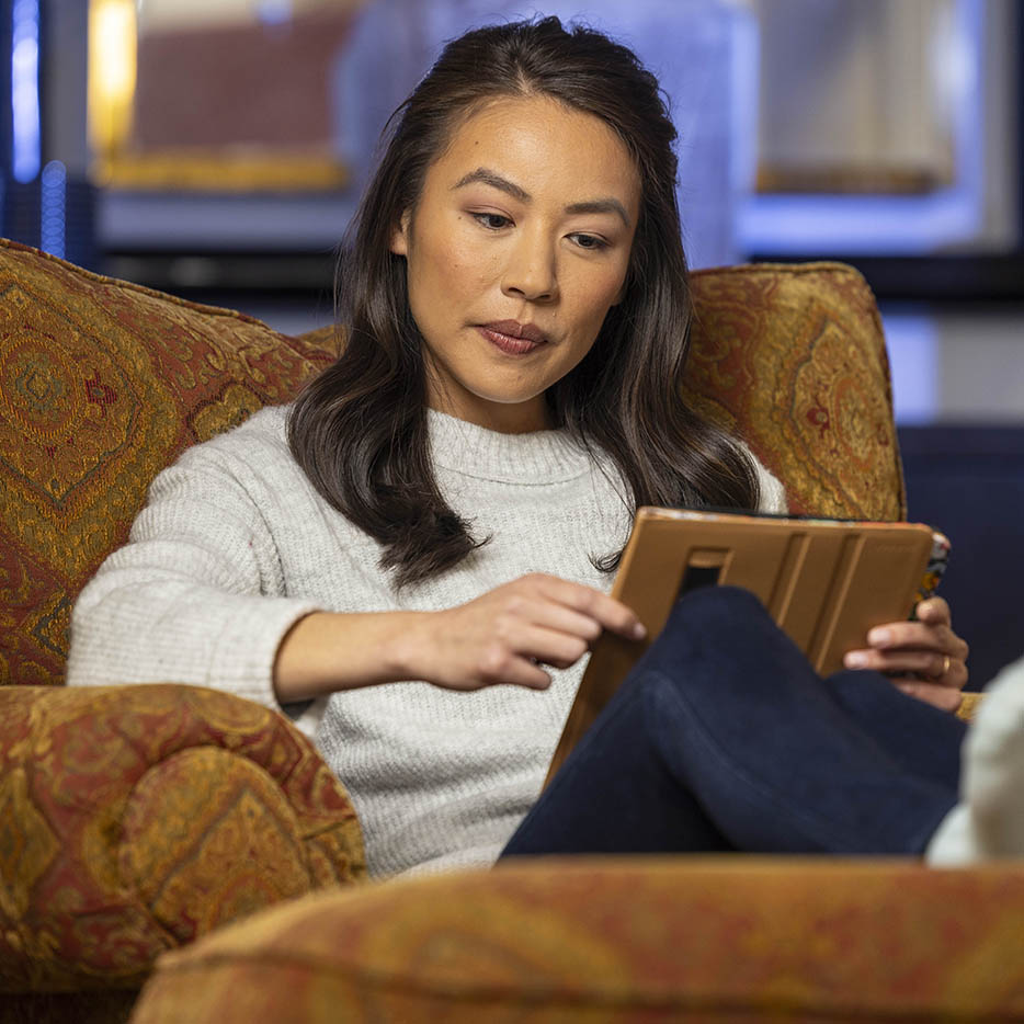 A woman with medium-length brown hair, wearing a cozy gray sweater, sits in an armchair, intently looking at a tablet in a brown case.