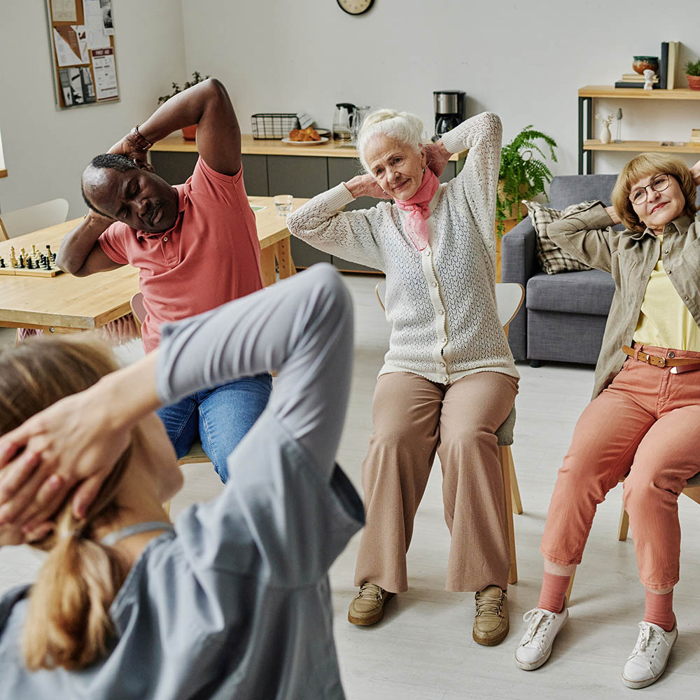 Three seniors sit on chairs in a bright room, following an instructor's lead by stretching their arms behind their heads during a group exercise class.