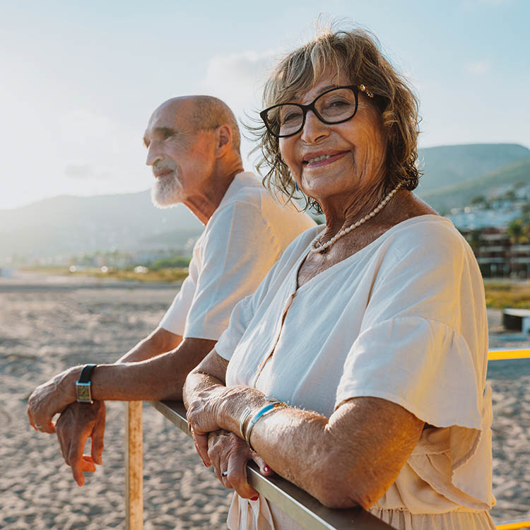 Smiling senior woman in glasses and a pearl necklace standing at a beach railing with her partner.