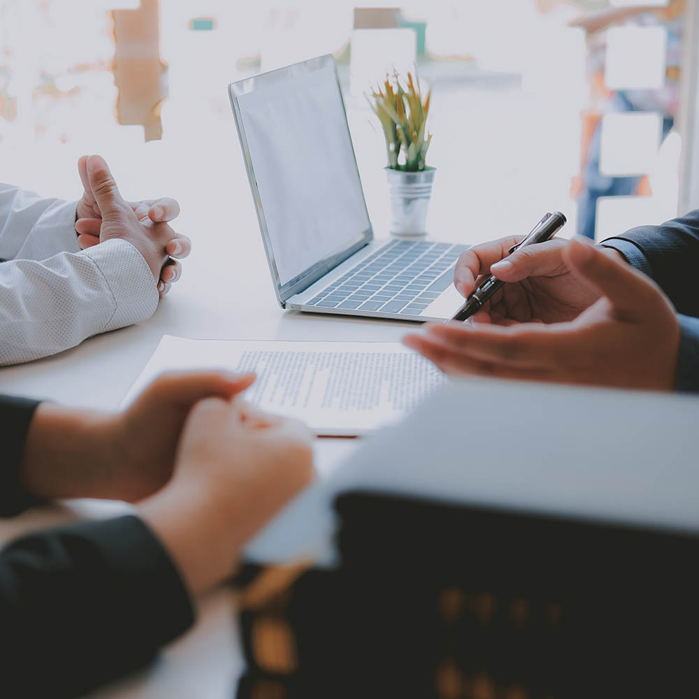 Close-up of hands over a desk with a laptop and documents, appearing to discuss a professional agreement or plan.