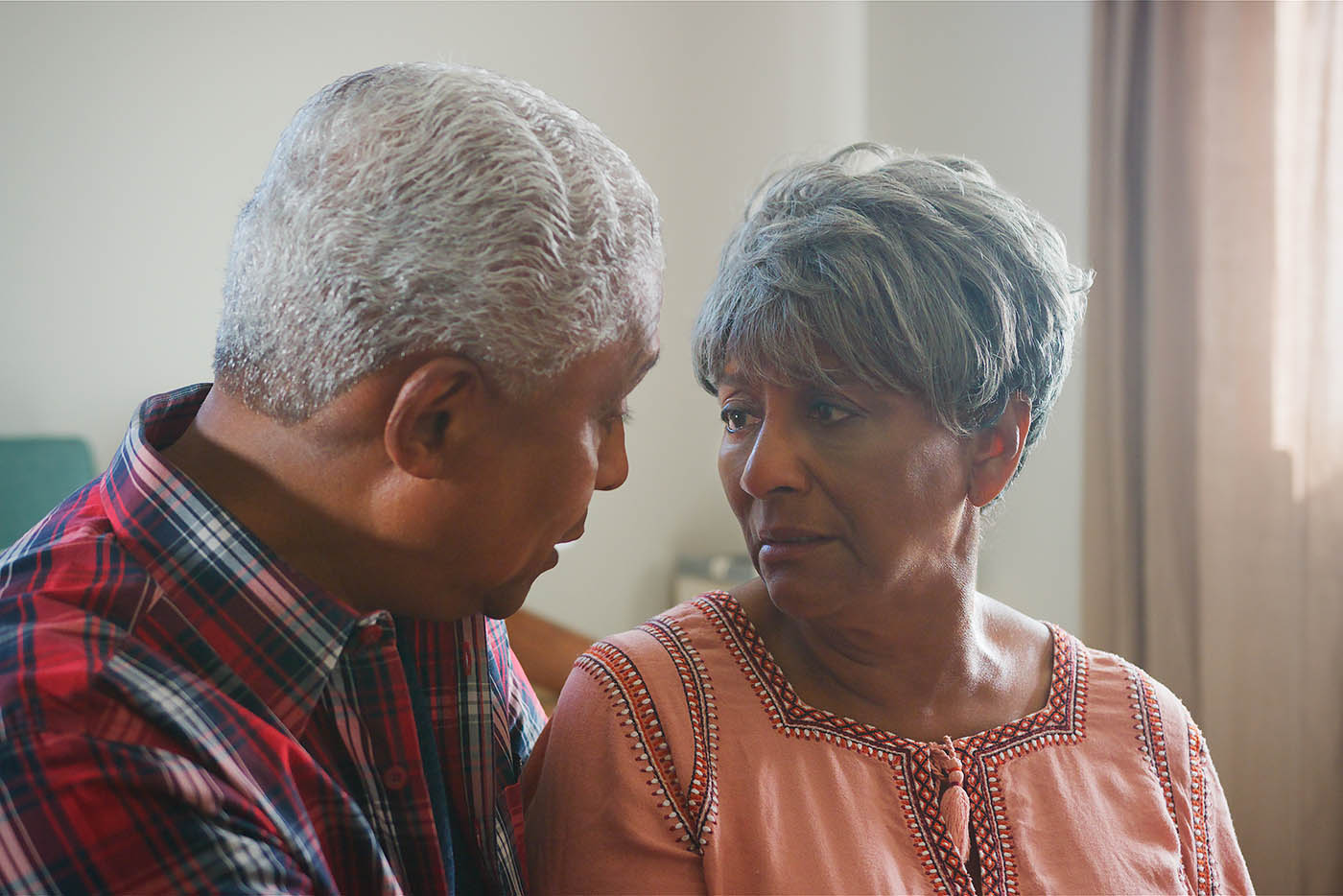 An elderly man in a plaid shirt leans in to speak softly to an elderly woman, who looks back with a serious and attentive expression.