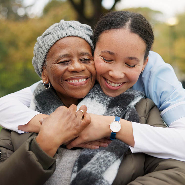 A young woman in a blue uniform smiling and embracing an elderly woman from behind outdoors.